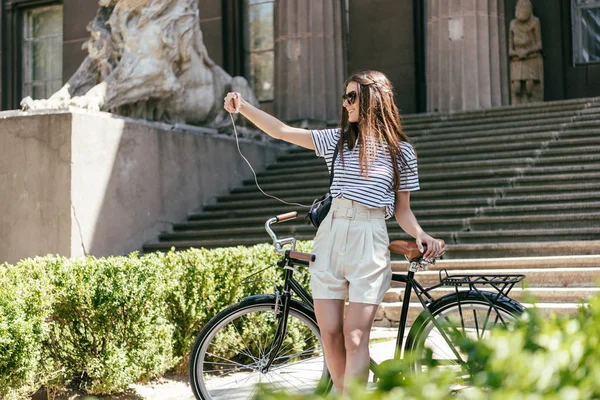 Happy young oman in sunglasses taking selfie with smartphone while standing with bicycle near beautiful building with columns and stairs — Stock Photo