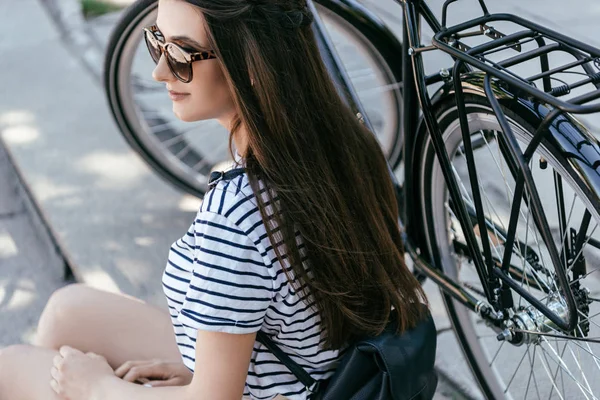 Высокий угол зрения привлекательной девушки в солнечных очках, глядя в сторону, сидя на лестнице рядом с велосипедом — стоковое фото