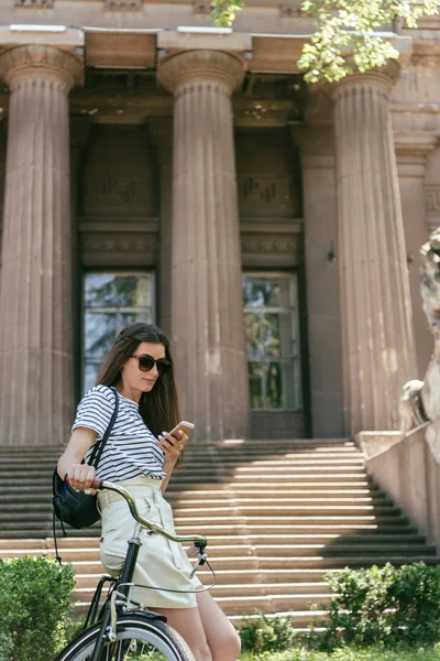 Attractive girl in sunglasses sitting on bike and using smartphone near beautiful building with columns and stairs — Stock Photo