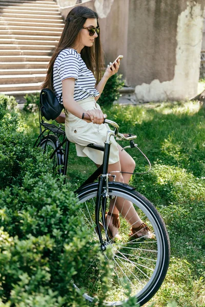 Красивая молодая женщина в солнечных очках сидит на велосипеде и использует смартфон — стоковое фото