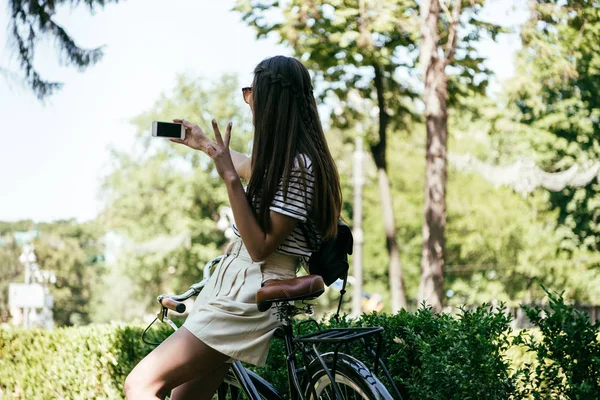 Beautiful girl sitting on bicycle and taking selfie with smartphone — Stock Photo