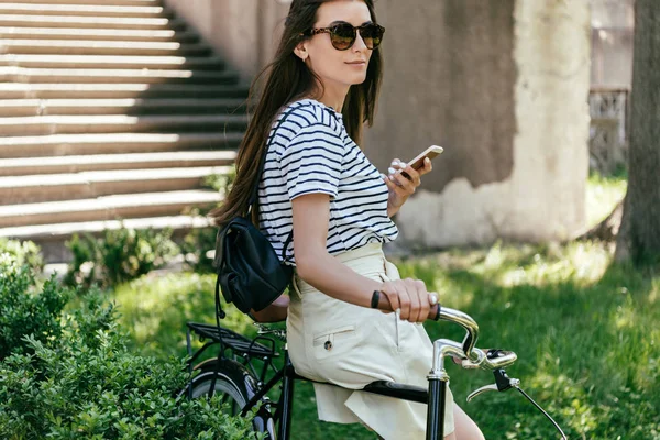 Красивая молодая женщина в солнечных очках сидит на велосипеде и использует смартфон на улице — стоковое фото