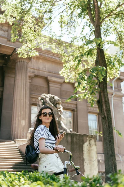 Schönes lächelndes Mädchen mit Sonnenbrille, das mit Fahrrad steht und Smartphone benutzt — Stockfoto