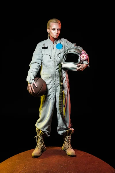 Cosmonauta en traje espacial sosteniendo pelota de baloncesto y casco con flor mientras está de pie en el planeta rojo - foto de stock