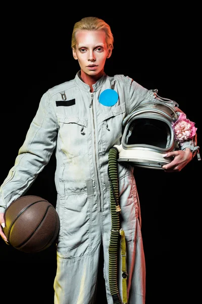 Atractiva astronauta hembra en traje espacial sosteniendo baloncesto y casco con flor, aislado en negro - foto de stock