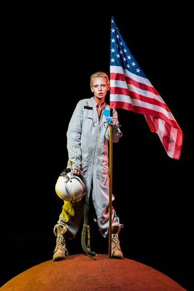 Atractiva astronauta femenina en traje espacial sosteniendo casco y bandera americana en el planeta - foto de stock