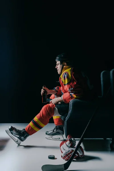 Side view of emotional professional hockey player drinking beer and watching tv on black — Stock Photo