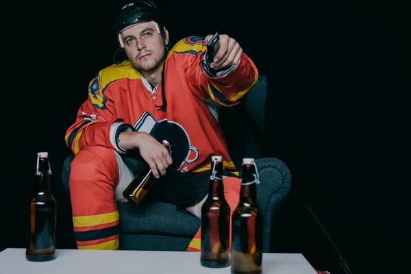Hockey player holding beer bottle and using remote controller while watching tv on black — Stock Photo