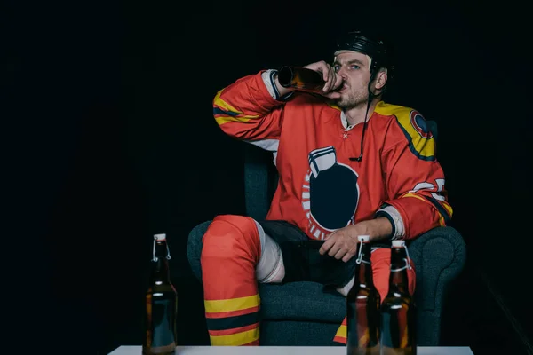 Jeune joueur de hockey boire de la bière tout en étant assis dans le fauteuil et regarder la télévision sur noir — Photo de stock