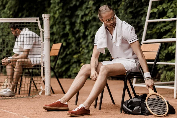 Retro styled tennis player sitting on chair with towel and putting racket on bag at court — Stock Photo