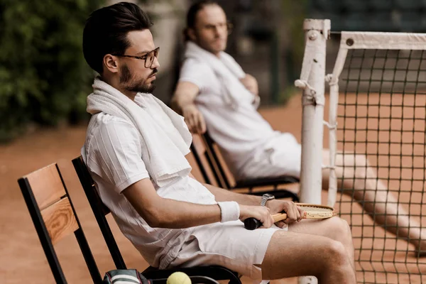 Vista lateral de jugadores de tenis de estilo retro que descansan en sillas con toallas y raquetas en la cancha de tenis - foto de stock