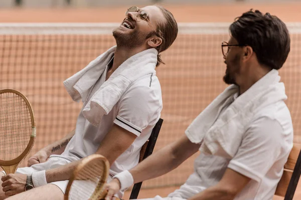 Joueurs de tennis souriants de style rétro assis sur des chaises avec des serviettes et des raquettes au court de tennis — Photo de stock