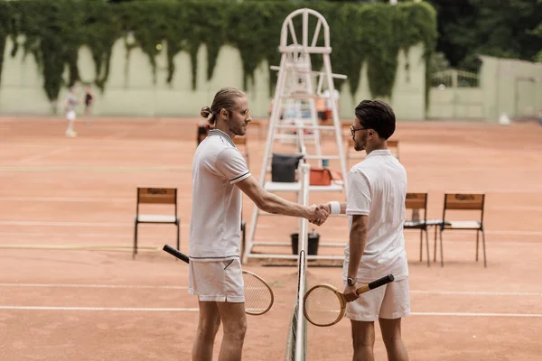 Vista lateral de jugadores de tenis de estilo retro estrechando la mano por encima de la red de tenis en la cancha - foto de stock