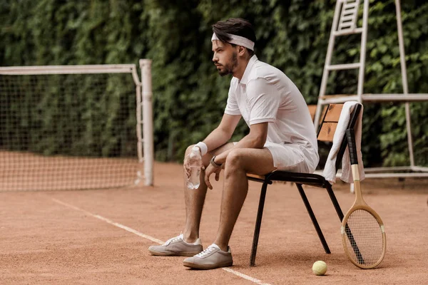 Вид сбоку красивого теннисиста в стиле ретро, сидящего на стуле с бутылкой воды на теннисном корте — стоковое фото