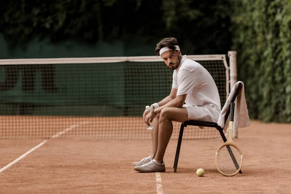 Красивый теннисист в стиле ретро сидит на стуле с бутылкой воды на теннисном корте — стоковое фото