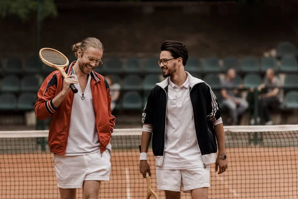 Lächelnde Tennisspieler im Retro-Stil, die mit Schlägern am Tennisplatz spazieren gehen — Stockfoto