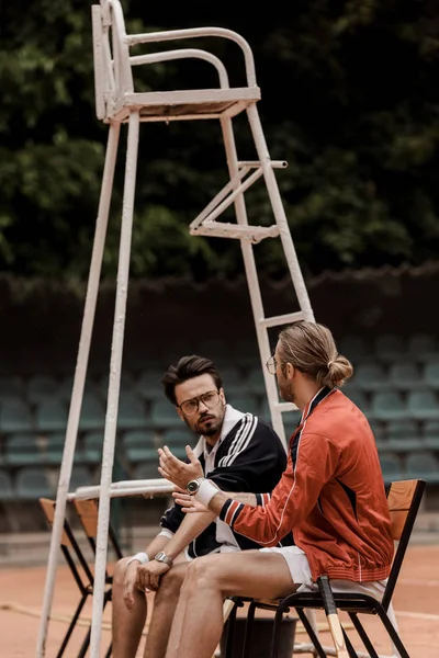 Jugadores de tenis de estilo retro hablando y sentado en sillas en la cancha de tenis - foto de stock