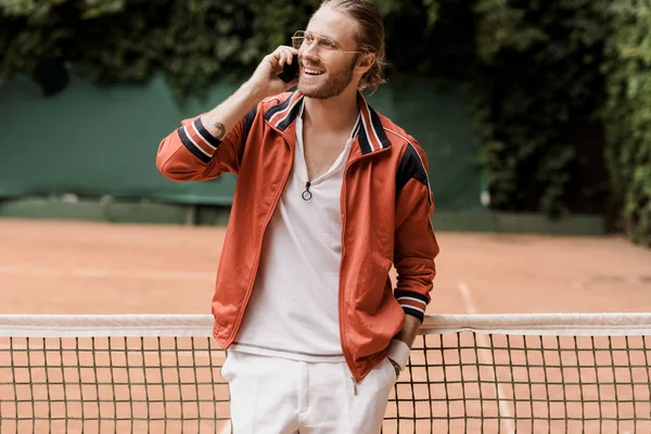 Sonriente jugador de tenis estilo retro hablando por teléfono inteligente en la cancha de tenis - foto de stock