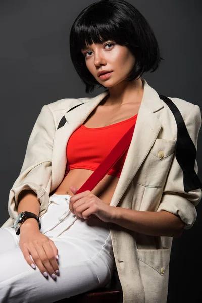 Beautiful young woman in stylish jacket and untied bowtie sitting on chair isolated on grey — Stock Photo