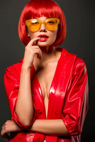 Retrato de cerca de la hermosa mujer joven en chaqueta de látex rojo en piel desnuda y gafas de sol de color amarillo vintage mirando a la cámara aislada en gris - foto de stock