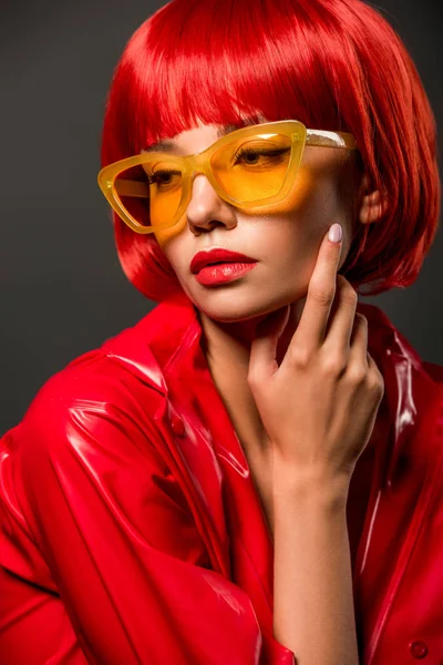 Retrato de cerca de la mujer joven de moda en chaqueta de látex rojo y gafas de sol de color amarillo vintage aisladas en gris - foto de stock