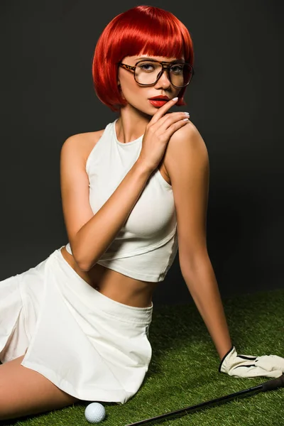 Seductive young woman with red bob cut sitting on green grass with golf equipment and looking at camera on grey — Stock Photo