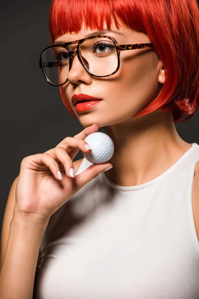 Close-up portrait of attractive young woman with red bob cut and stylish eyeglasses holding golf ball isolated on grey — Stock Photo