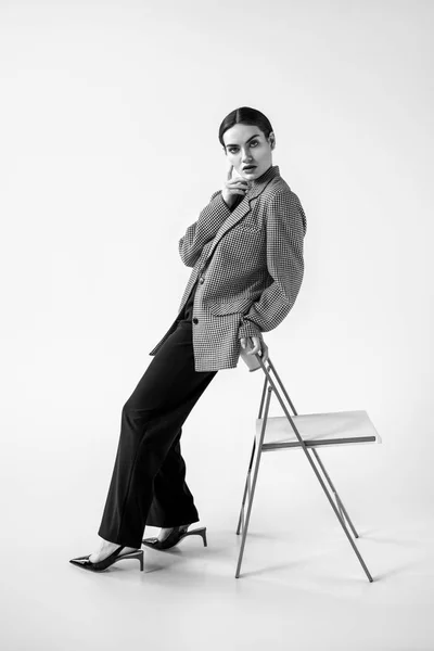 Beautiful glamour model posing in fashionable vintage suit near chair, on grey, black and white — Stock Photo