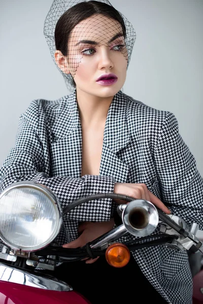 Mujer elegante en chaqueta y velo neto posando en scooter clásico retro, aislado en gris - foto de stock