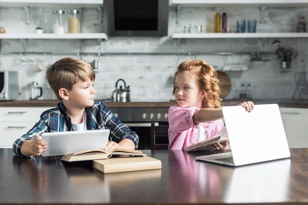 Hermano pequeño y hermana con dispositivos digitales sentados en la cocina y mirándose unos a otros - foto de stock