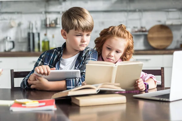 Concentrated little brother and sister with devices reading book together — Stock Photo