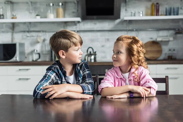 Hermano pequeño y hermana sentados en la cocina juntos y mirándose el uno al otro - foto de stock