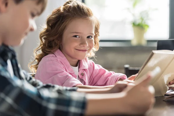 Entzückende kleine Schülerin schaut in die Kamera, während sie mit Bruder Buch liest, Tablet im Vordergrund verschwommen — Stockfoto