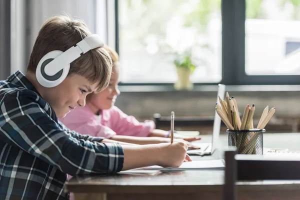 Вид збоку усміхненого маленького школяра малюнок з кольоровими олівцями, а його сестра використовує ноутбук — стокове фото
