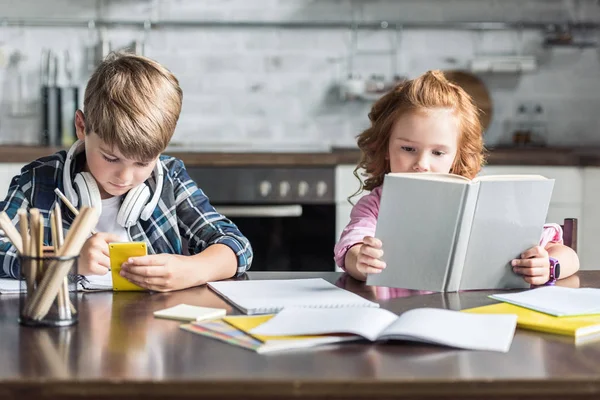 Сосредоточенные маленькие братья и сестры делают домашнюю работу вместе на кухне — стоковое фото