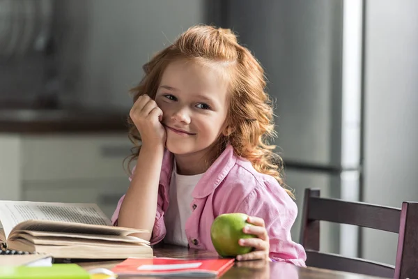 Nettes kleines Schulmädchen mit grünem Apfel schaut während der Hausaufgaben in die Kamera — Stockfoto