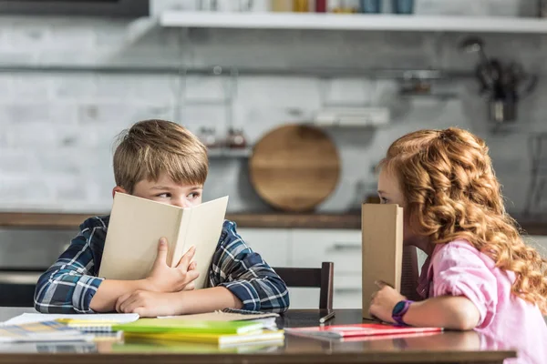 Hermano pequeño y hermana cubriendo caras con libros y mirándose - foto de stock