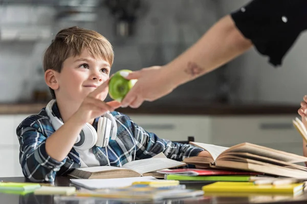 Recortado disparo de madre dando verde manzana a su hijo mientras él haciendo tarea - foto de stock