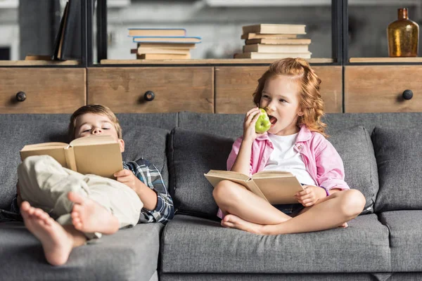 Lindo hermanito y hermana leyendo libros en el sofá en casa - foto de stock