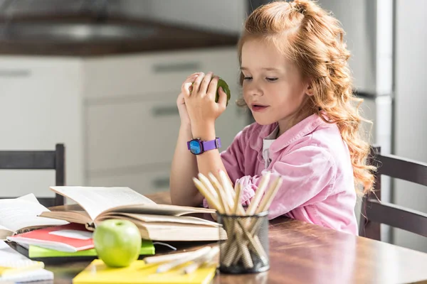 Cute little schoolgirl with green apple doing homework — Stock Photo