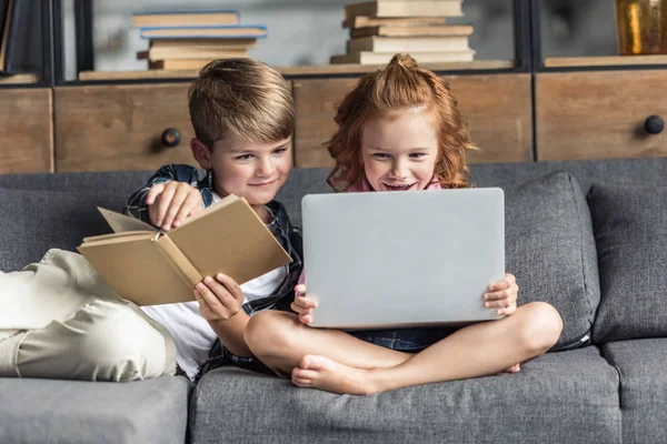 Маленький брат і сестра використовують ноутбук і читання книги, розслабляючись на дивані — стокове фото