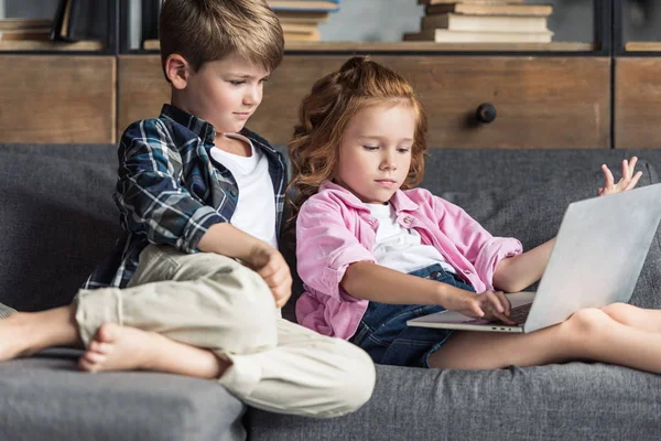 Маленький брат і сестра використовують ноутбук разом, розслабляючись на дивані — стокове фото
