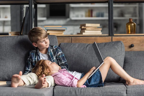 Mignonne petite fille couchée avec la tête sur les jambes du frère et en utilisant un ordinateur portable pendant qu'ils se détendent sur le canapé — Photo de stock