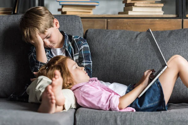 Little girl lying with head on legs of brother and using laptop while they relaxing on couch — Stock Photo