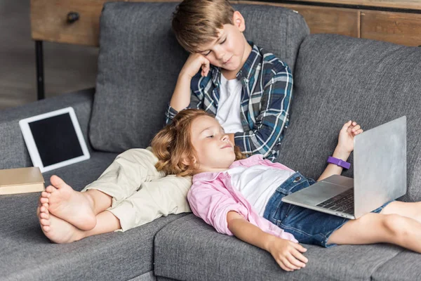 Little brother and sister relaxing on couch with gadgets — Stock Photo