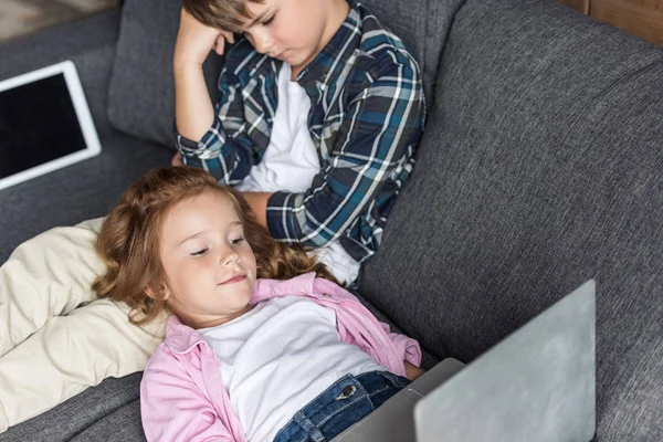 Vista de ángulo alto de hermano pequeño y hermana relajarse en el sofá con gadgets - foto de stock