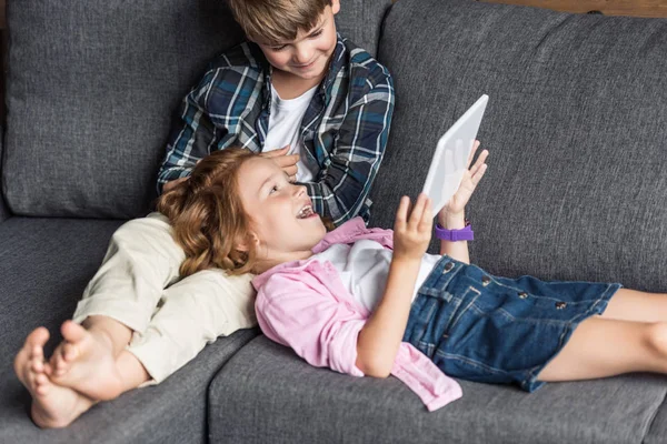 Младшие брат и сестра вместе за планшетом, расслабляясь на диване — стоковое фото