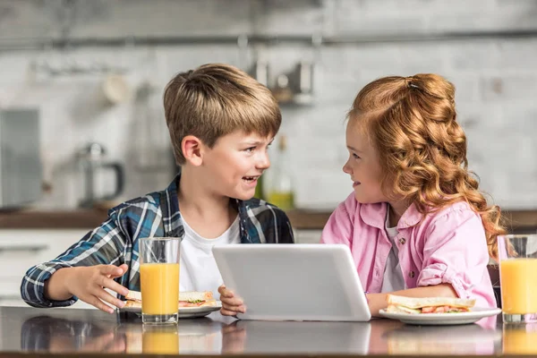 Felice fratellino e sorella utilizzando tablet durante la colazione e guardarsi l'un l'altro — Foto stock