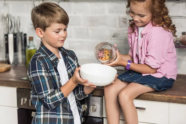 Adorable little brother and sister pouring macaroni into bowl from jar — Stock Photo