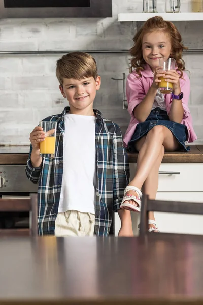 Irmãozinho e irmã com copos de suco de laranja na cozinha — Fotografia de Stock